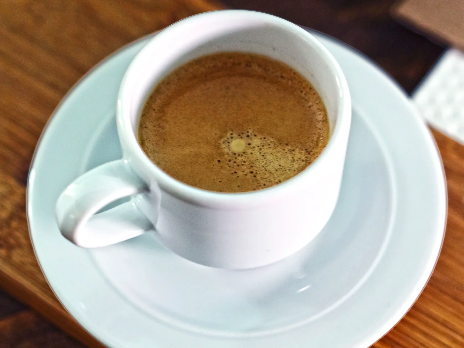 Xícara branca com café sobre um pires e uma mesa de madeira. Foto ilustrativa de Shameel Mukkath, Pexels.
