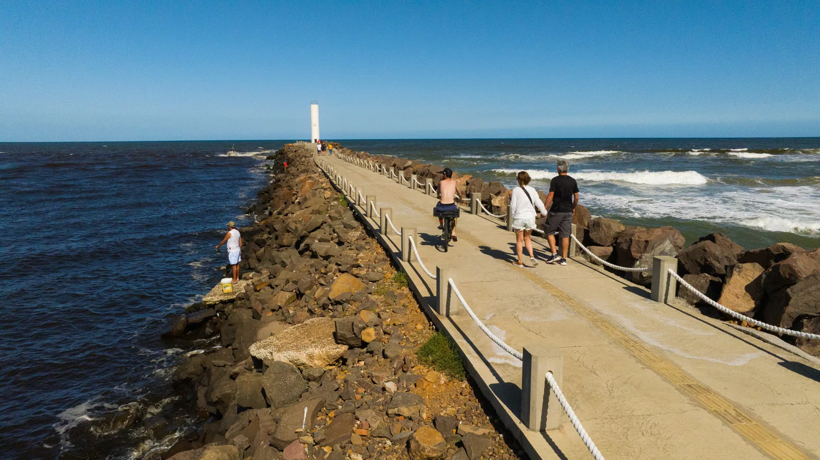Pessoas caminhando e pedalando pelo molhe, com o farol e o mar ao fundo