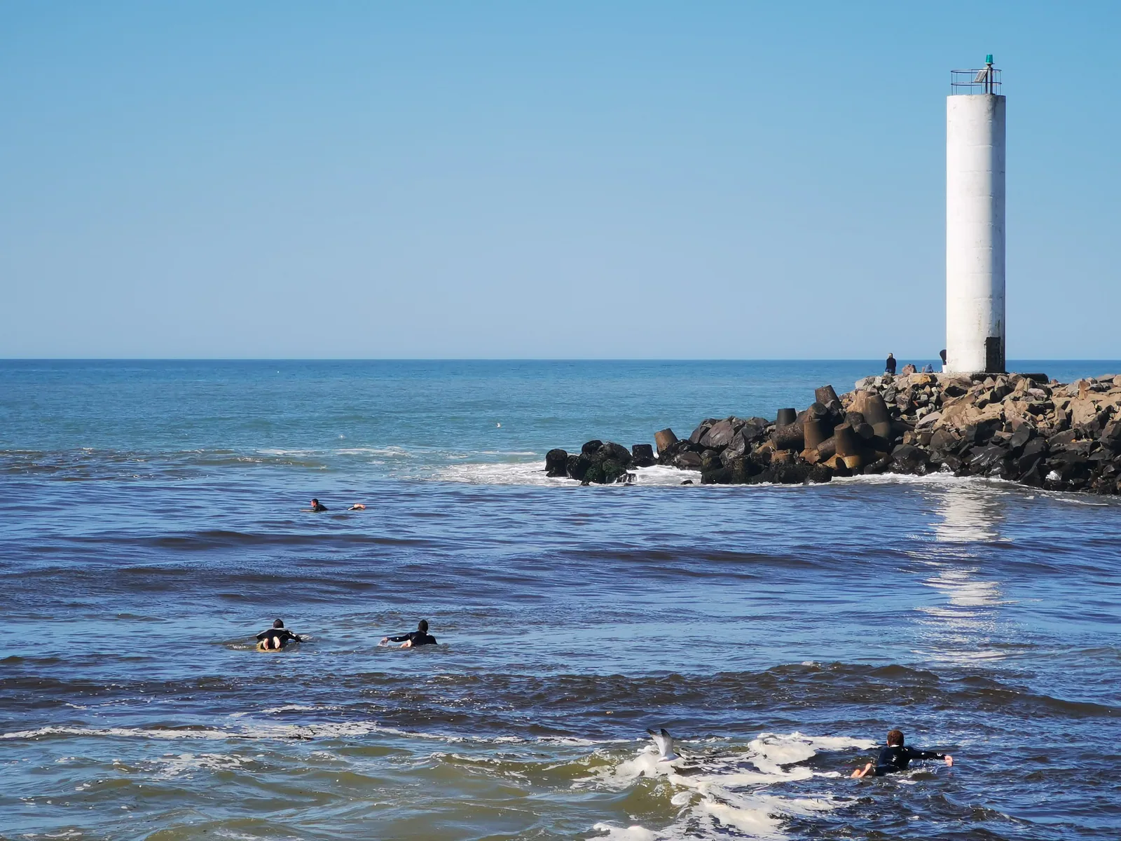 Surfistas no mar junto ao farol e às pedras dos Molhes da Barra