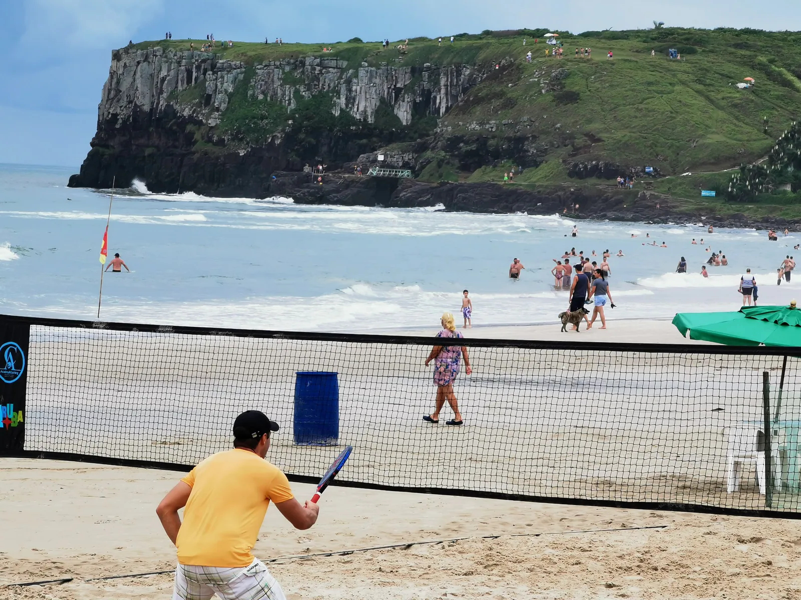 Jugador y red de tenis de playa en Praia da Cal, con bañistas y un acantilado al fondo