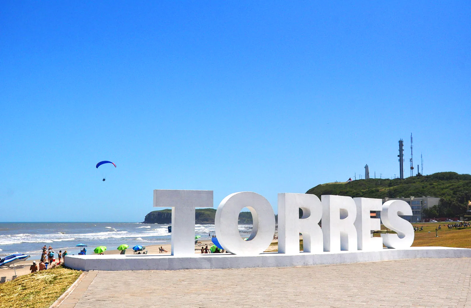Letreiro branco TORRES junto à Prainha, com o Morro do Farol e um parapente ao fundo