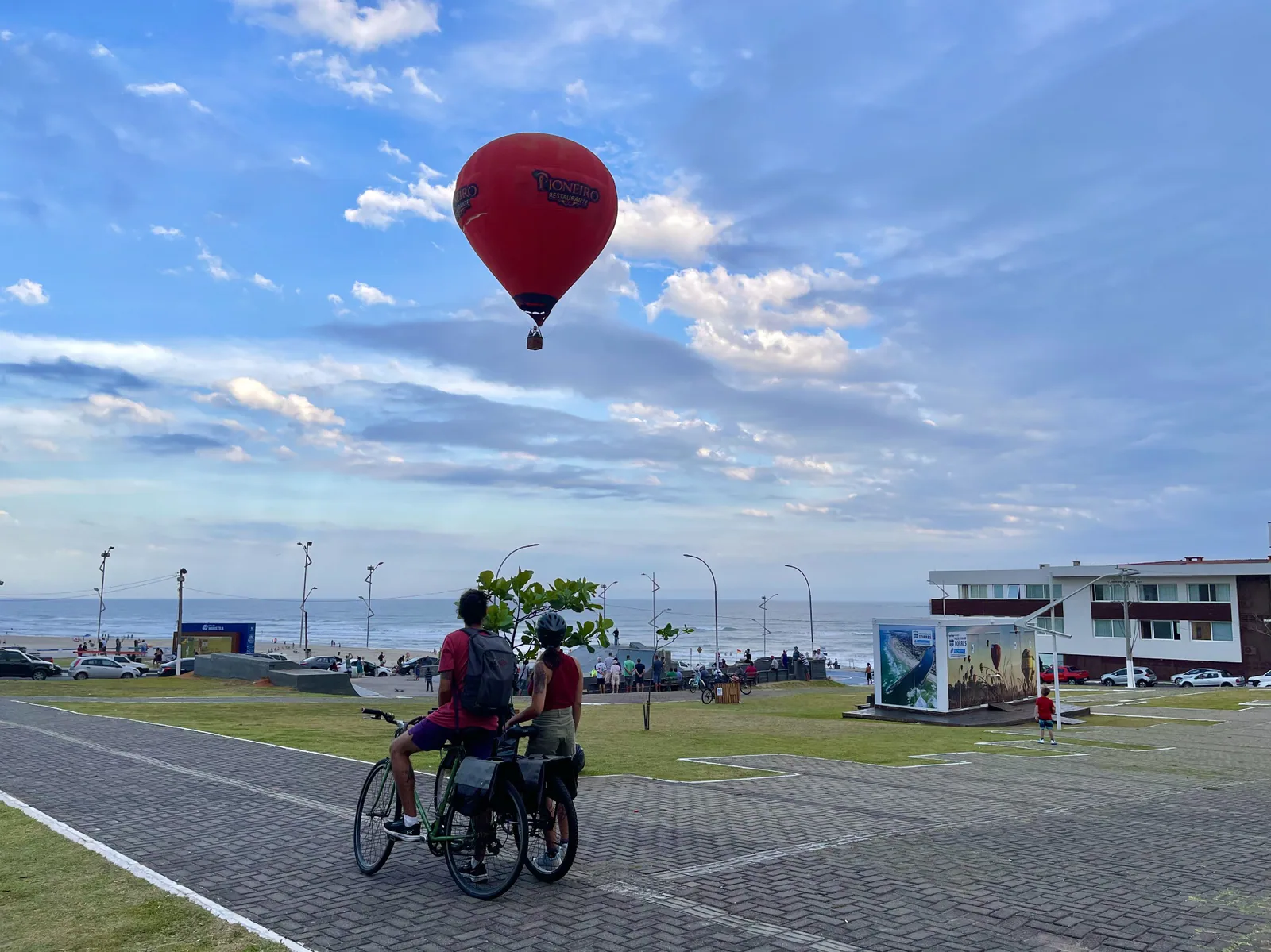 Balão sobre a Praça Pinheiro Machado, com ciclistas de costas, gramados e o mar ao fundo.
