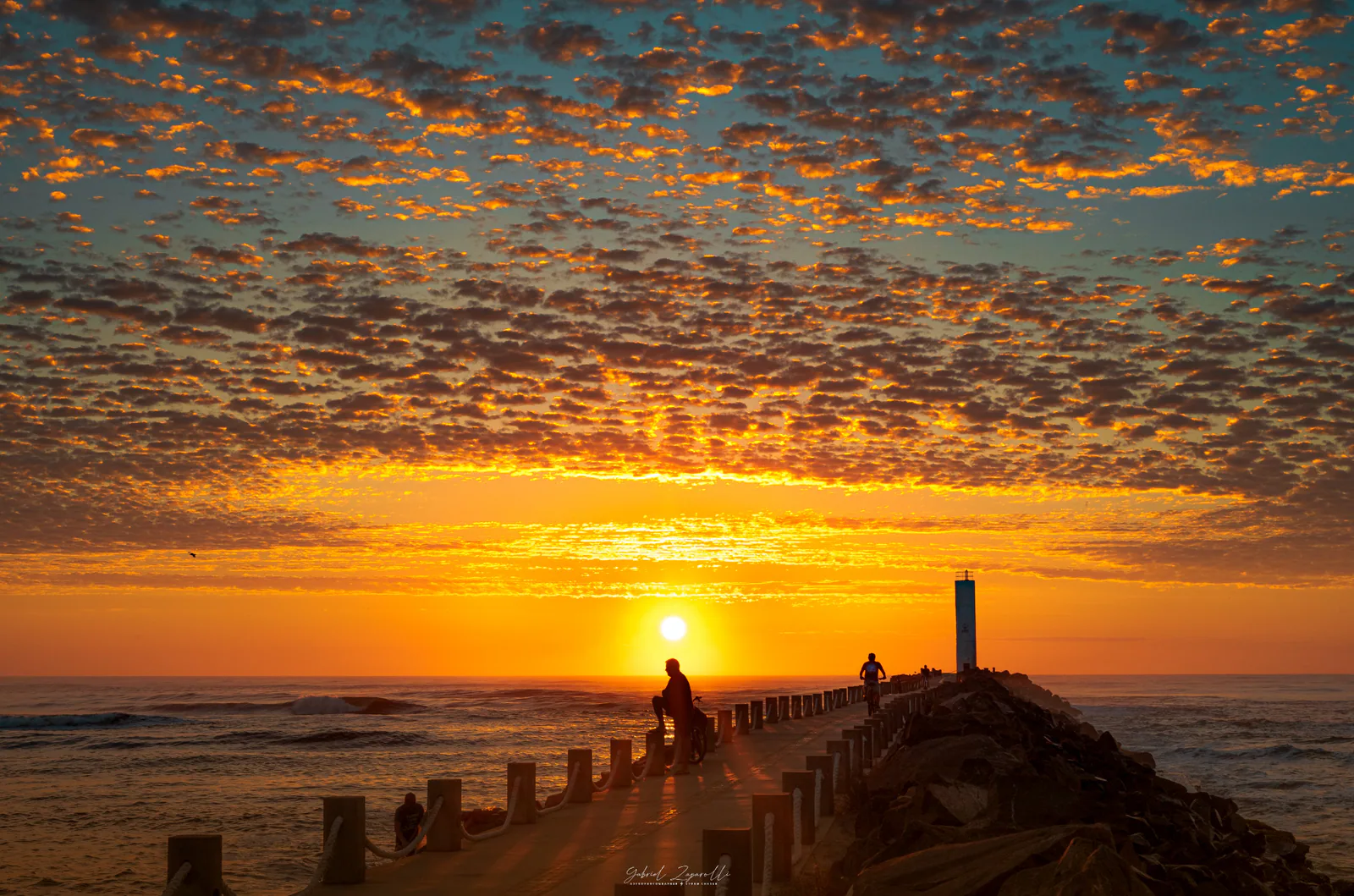 Caminho dos Molhes da Barra em direção ao farol, com o sol e nuvens iluminadas sobre o mar