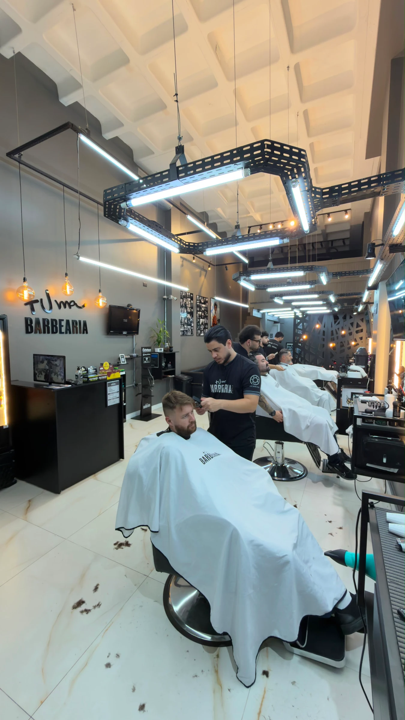 Barbeiros atendem clientes em cadeiras alinhadas no interior da Mais Uma Barbearia.